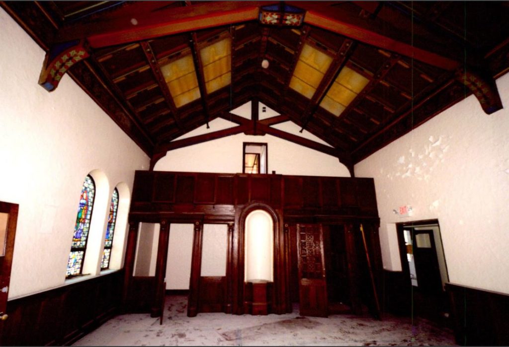 Inside of a chapel with exposed wood ceiling and rounded stained glass windows.