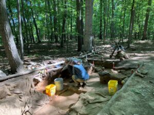 Two people crouching in a large dirt hole and surrounded by a forest.