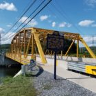 This image shows a yellow metal truss bridge with a blue and gold PA historical marker in front. The recently refurbished Bald Eagle's Nest marker is back in place in Centre County.