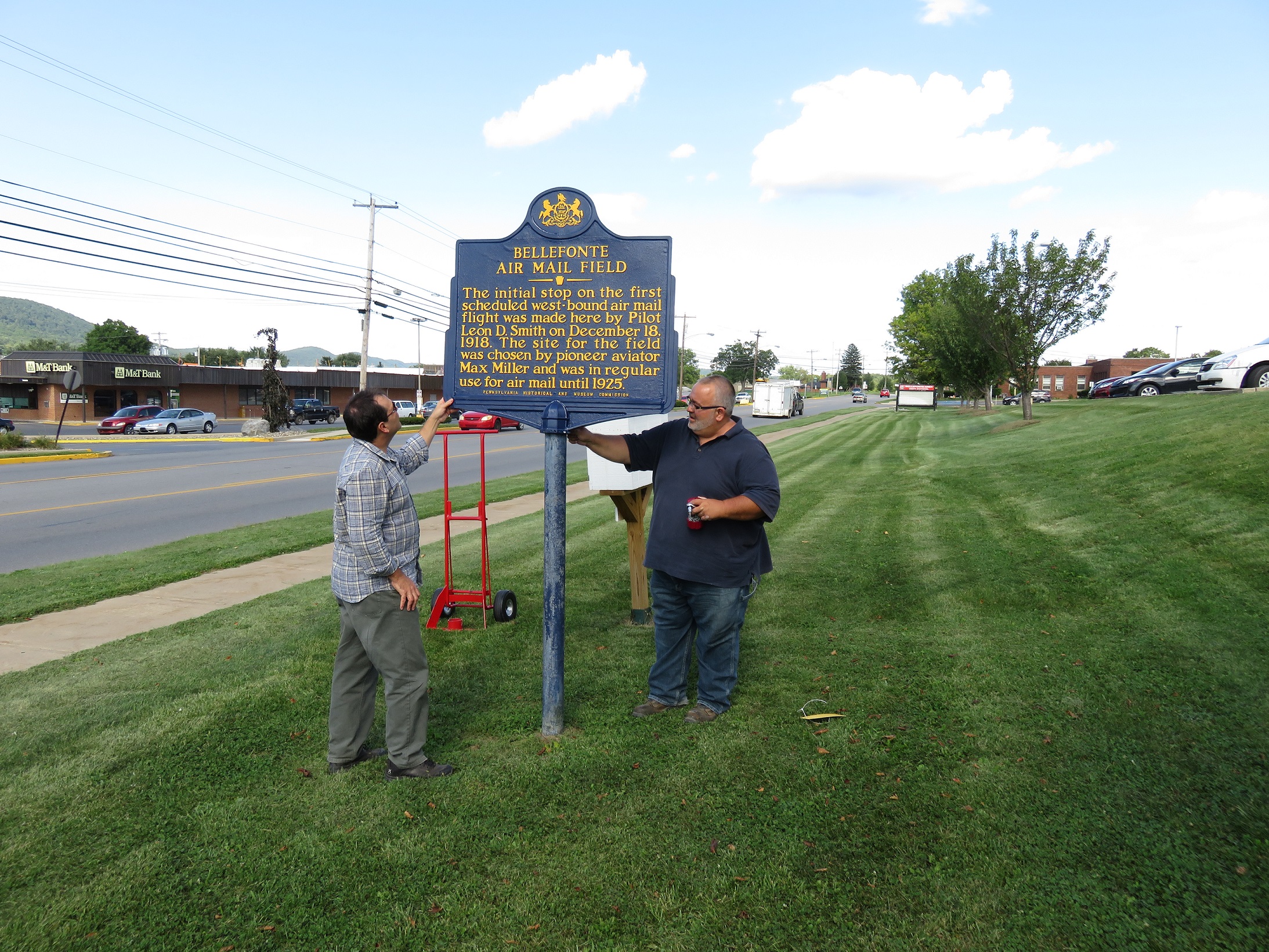 This image shows two men standing on a lawn holding a large blue and gold PA historical marker.
