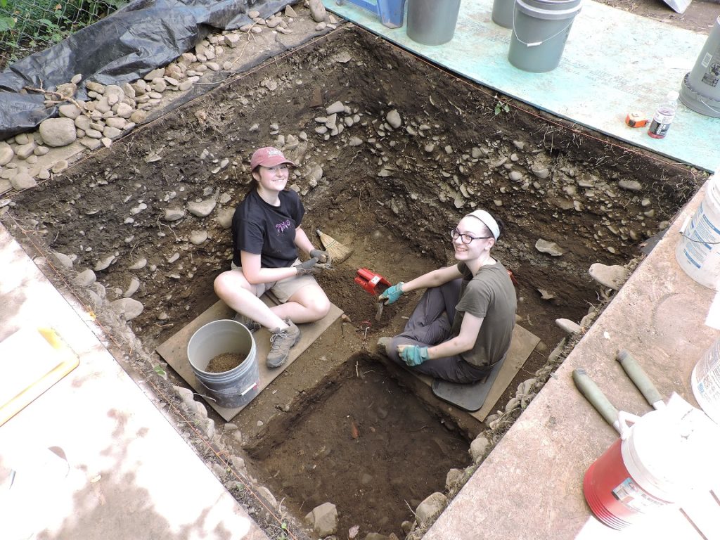 Two women sitting in a large, deep hole with trowels.