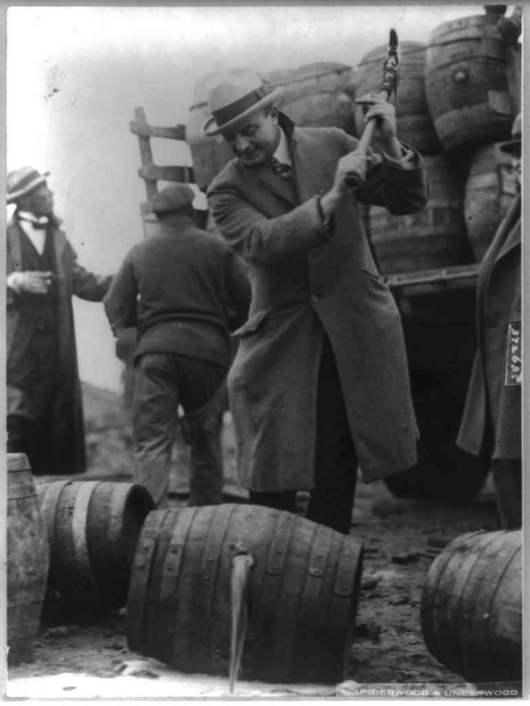 Man stands over barrel with axe. Liquid is pouring from barrel on the ground.  Truck full of barrels and other men in background.