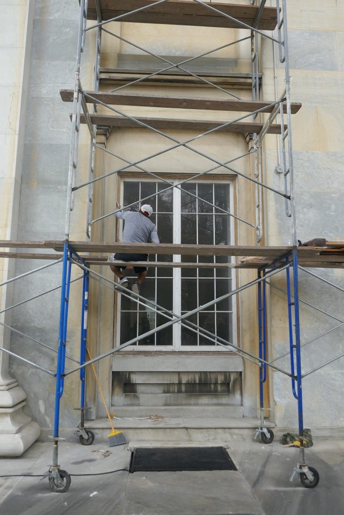 Man sitting on scaffolding working on tall wood window
