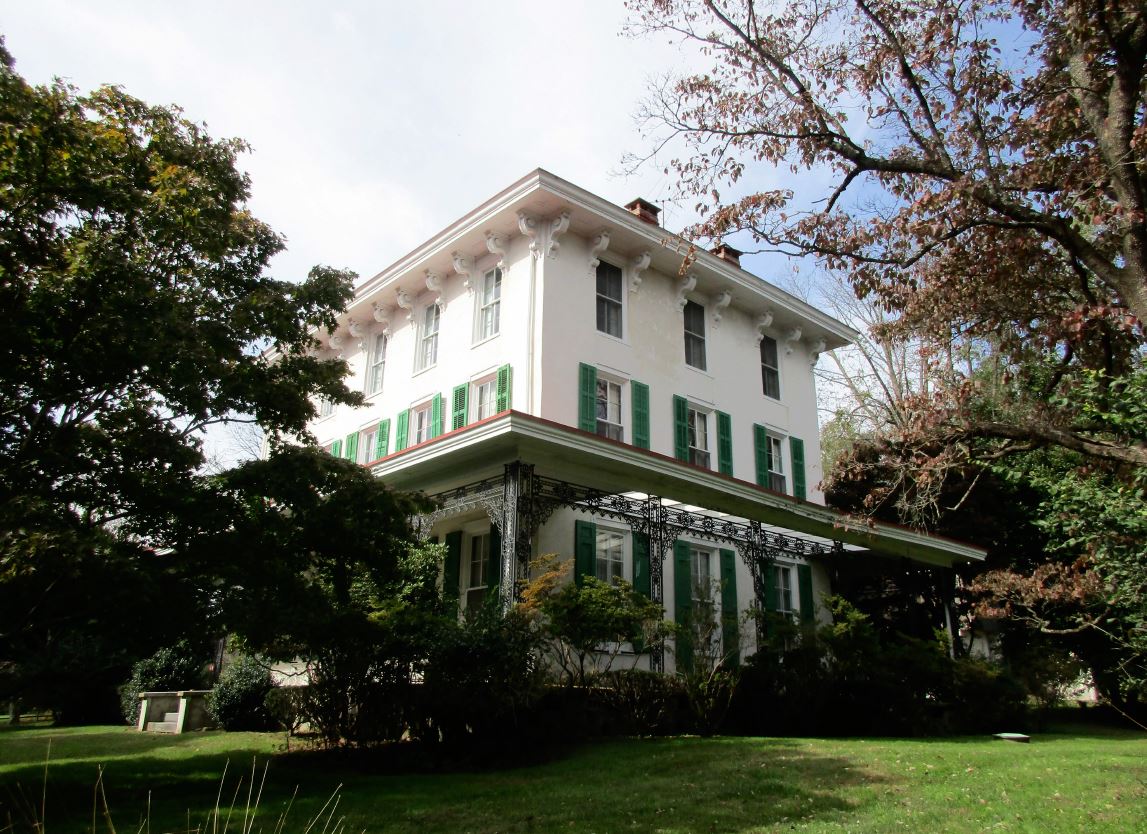 Large three story house with deep porch surrounded by grass and landscaping.