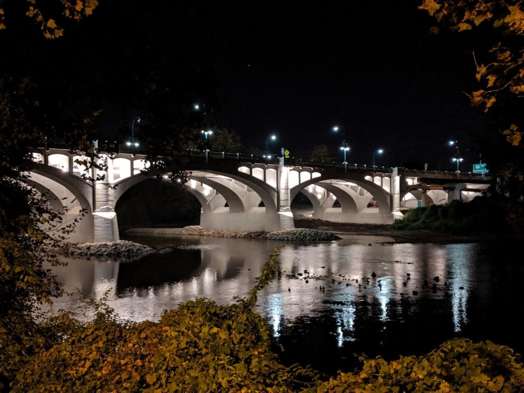 Looking across water at bridge at night with lights below spandrels.