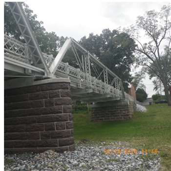 Looking at the side of a metal truss bridge on stone abutments over stone and grass.