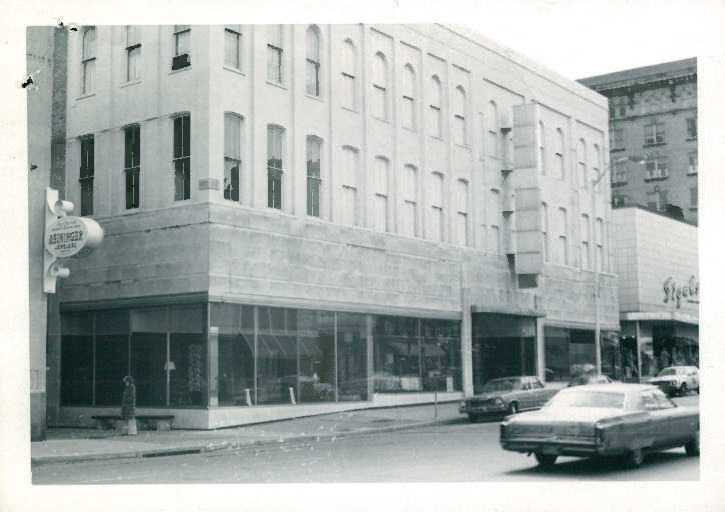 Black and white photograph of three-story building with large windows. Cars shown on street.
