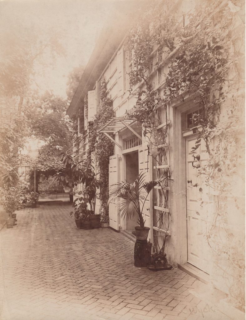 Sepia tone print of exterior wall with roses growing on a trellis and a brick walk.