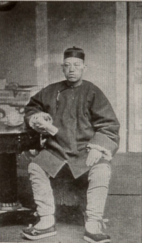 Black and white photograph of Chinese man in traditional dress sitting at a table.
