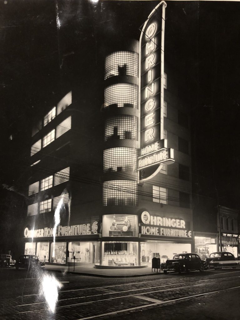 Black and white photograph of large retail store with neon sign light up at night.