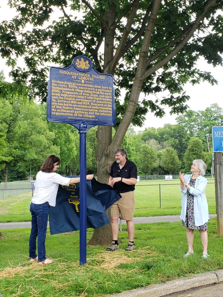 Two adults standing next to tall metal marker as third adult claps hands.