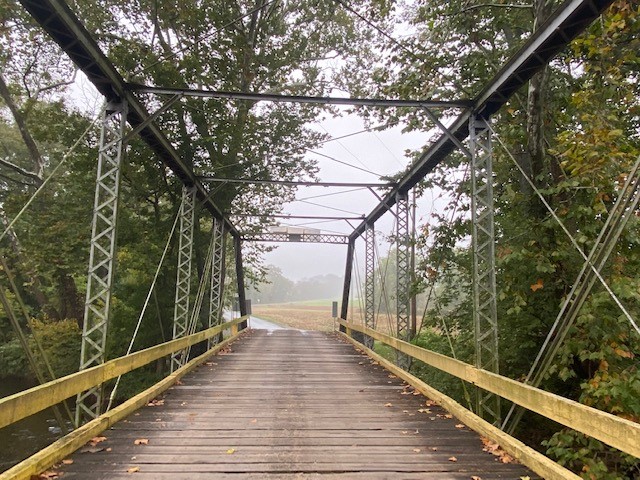Looking through metal truss bridge to road and fields beyond.