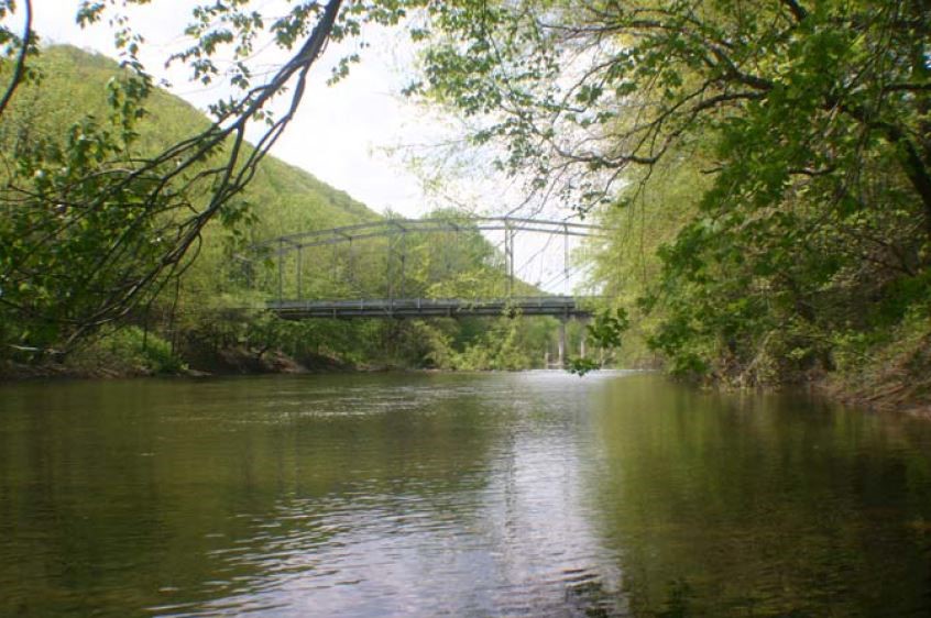 Metal truss bridge over wide stream with wooded areas on either side of stream.