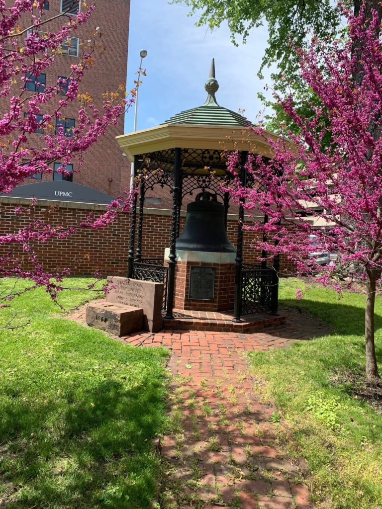 Brick and decorative iron gazebo surrounded by grass and flowering tress.