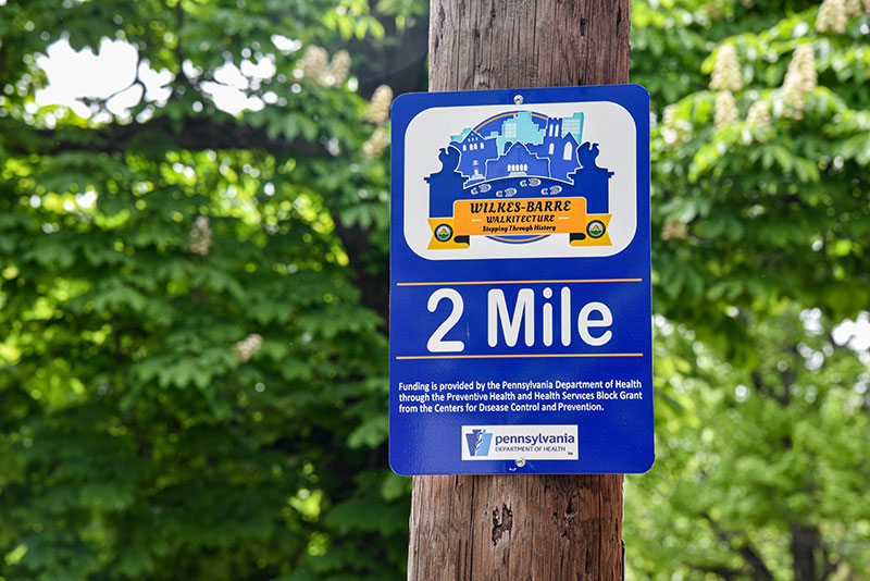 Small metal sign attached to a wood pole. Sign gives 2 mile mark and includes logo.
