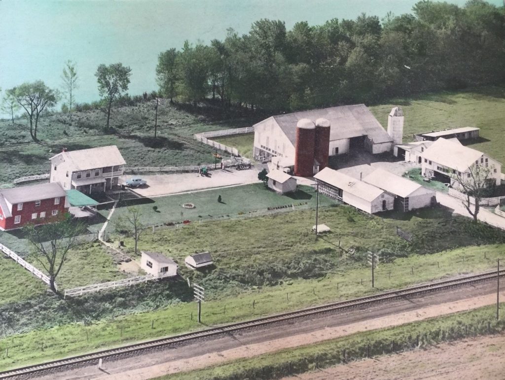 Aerial photograph of farm property. River at top of image and train tracks at botom. Large barn and outbuildings at right side of photograph and house at left.