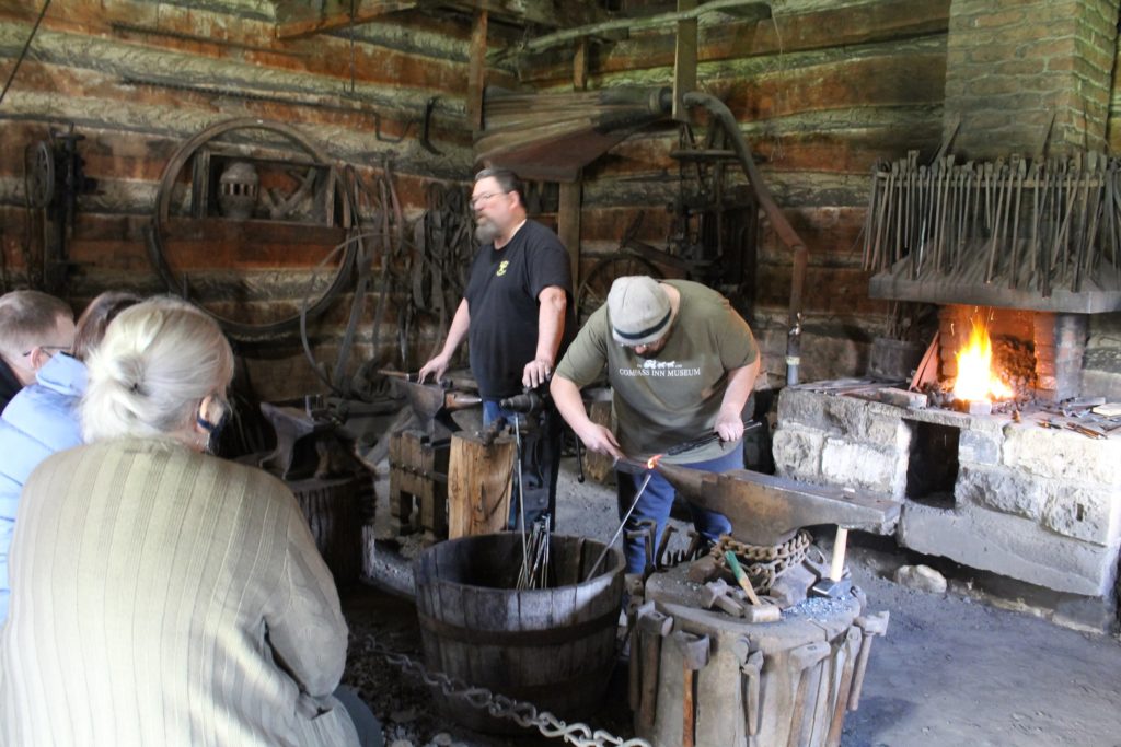 Two men working at on a large piece of metal in front of a seated people.