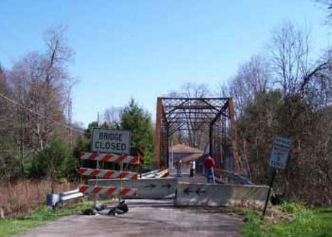 Signs and concrete barriers in front of a metal truss bridge.