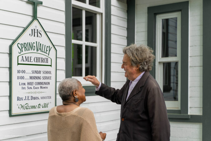 Two people stand in front of a building referencing a sign.