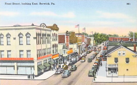 Historic color postcard of street with buildings on each side and parked cars.
