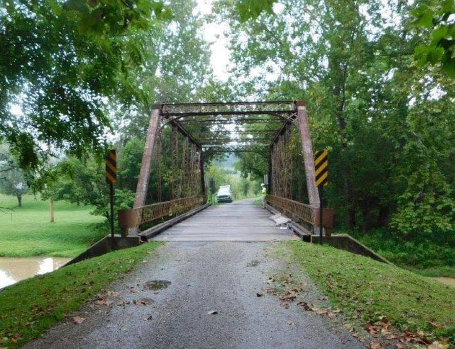 Road across a metal truss bridge, which is over water and surrounded by green trees.