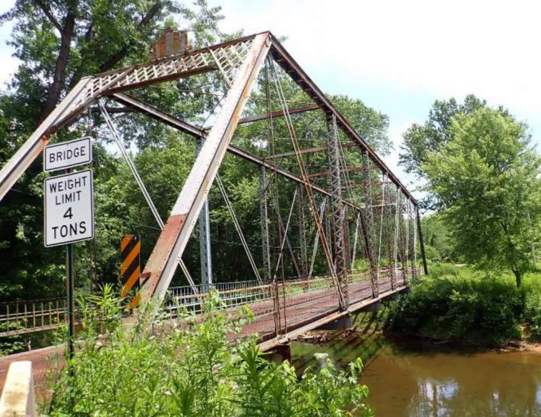 Metal truss bridge over water surrounded by green bushes and trees.