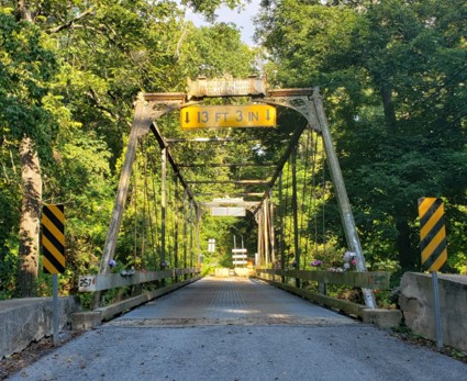 Road over a metal truss bridge that is surrounded by green trees.