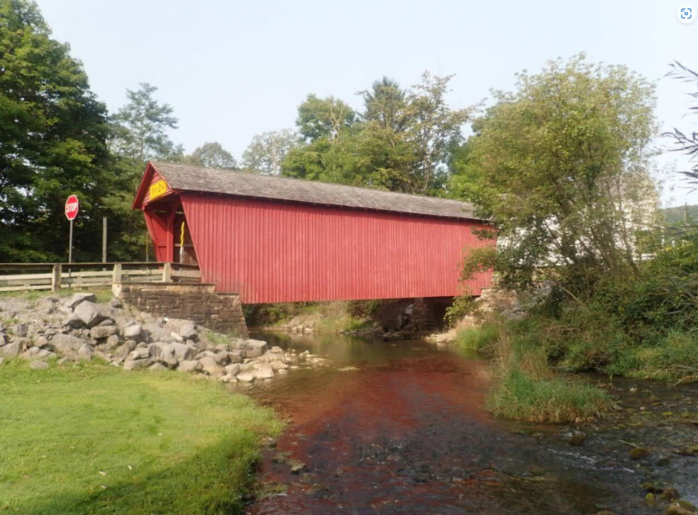 Covered bridge with vertical red siding and gable roof over a shallow creek.