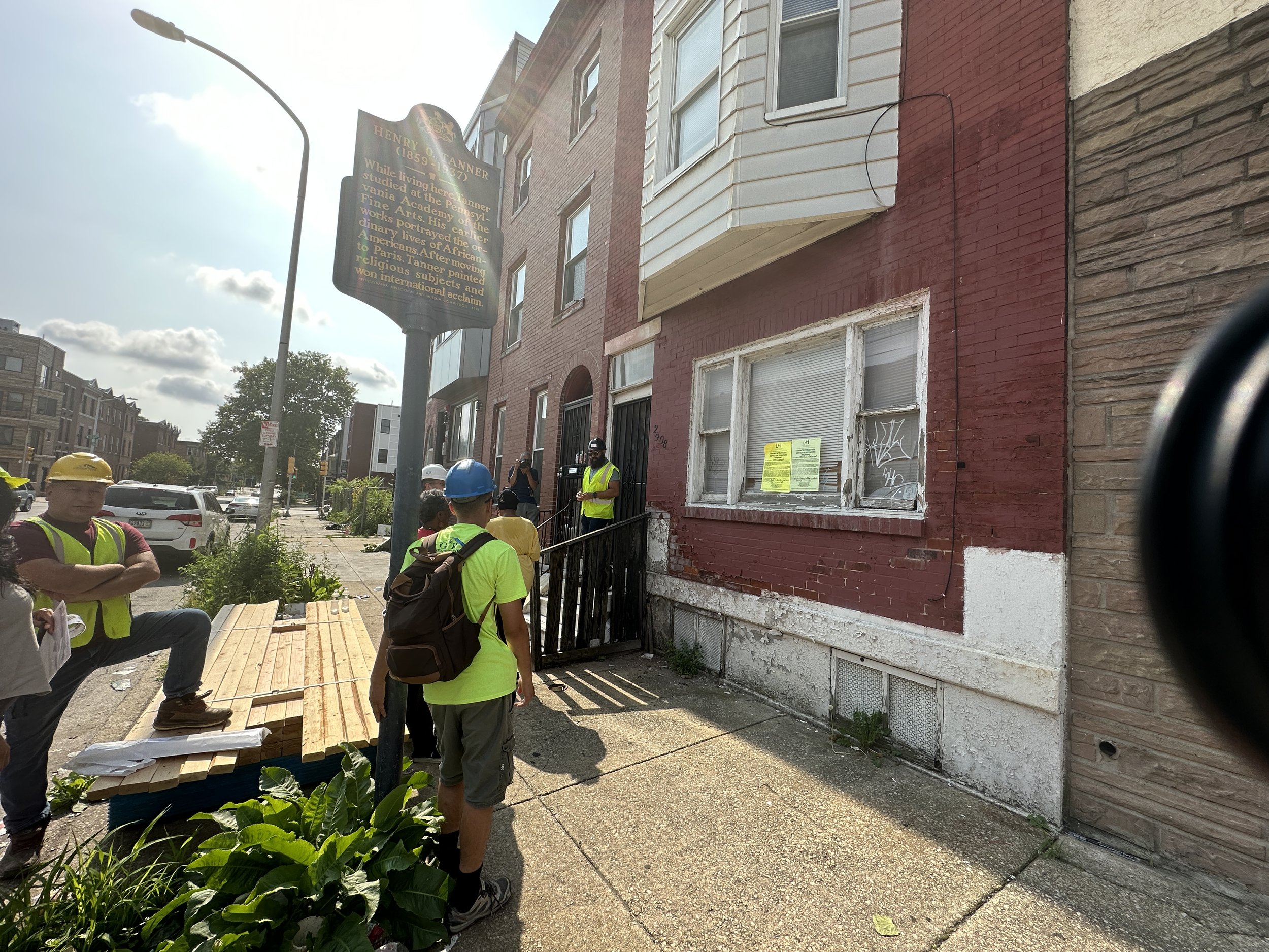 Group of people in hard hats and bright clothing on the sidewalk in front of a brick rowhouse.
