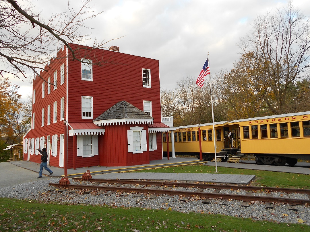 Large 3-story red building with white trim between rail tracks and a train and surrounded by grass.