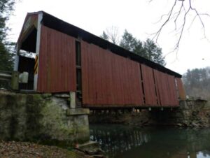 Covered bridge over water, set on masonry abutments and missing some vertical board siding.