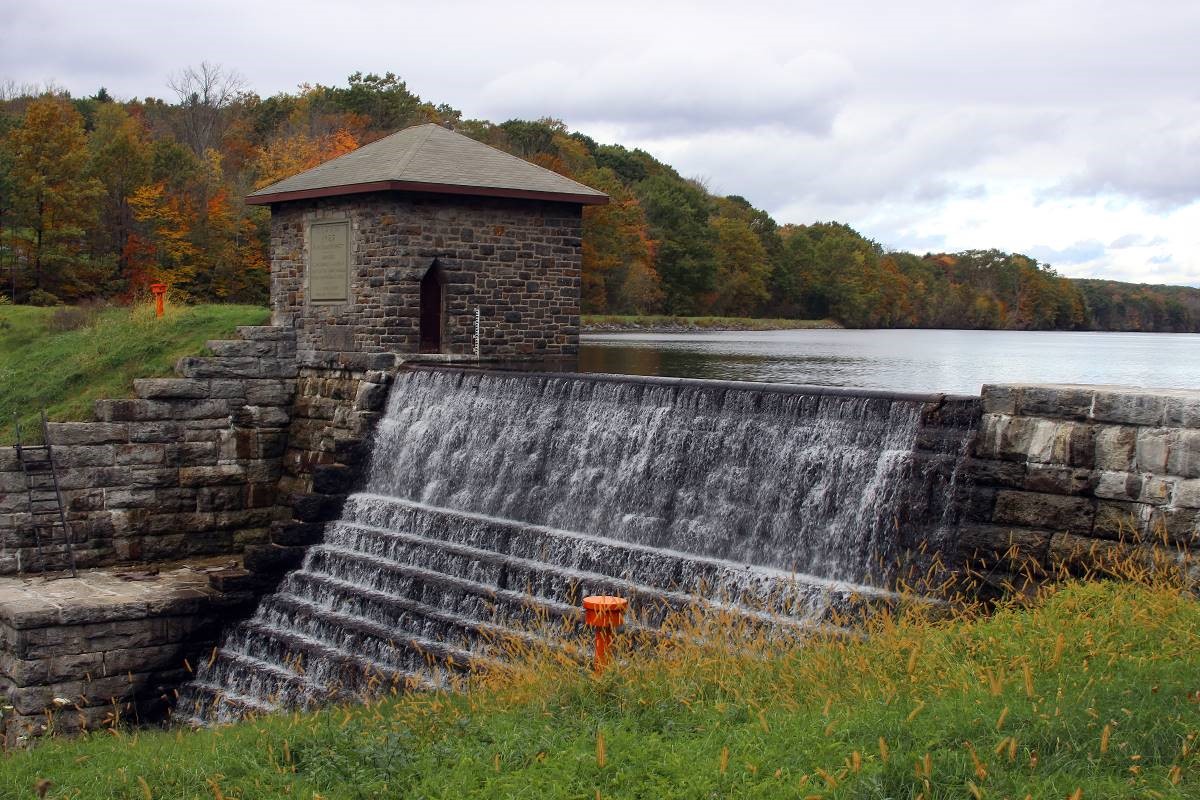 Water cascades down a stone dam from a body of water. A small one-story stone building stands adjacent to the water.