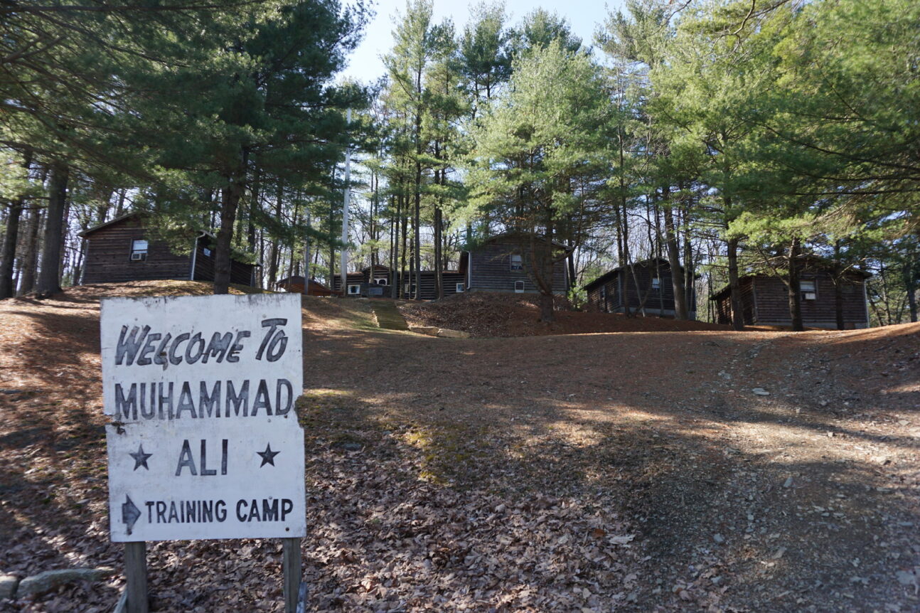Small white signs with the words "Welcome to Muhammed Ali training camp" in front of wooded area with several small log buildings.
