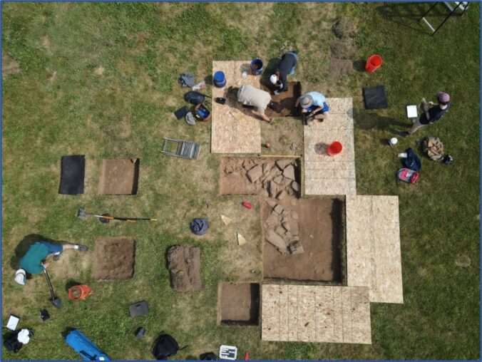 Aerial view of archaeology dig with excavation squares, exposed stones, plywood borders, and people and tools.