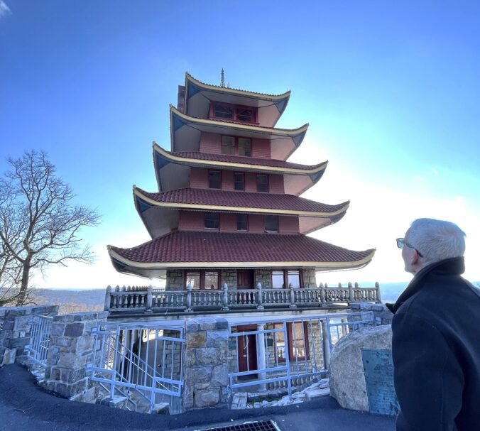 Man looking at large pagoda with many tiled roofs, stone foundation, and wood railing.
