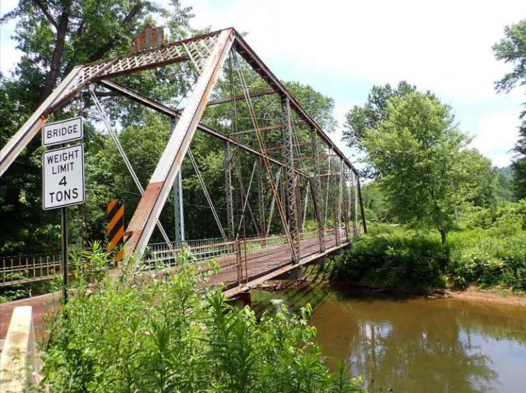 Metal truss bridge over shallow water with wooded and area at far end.