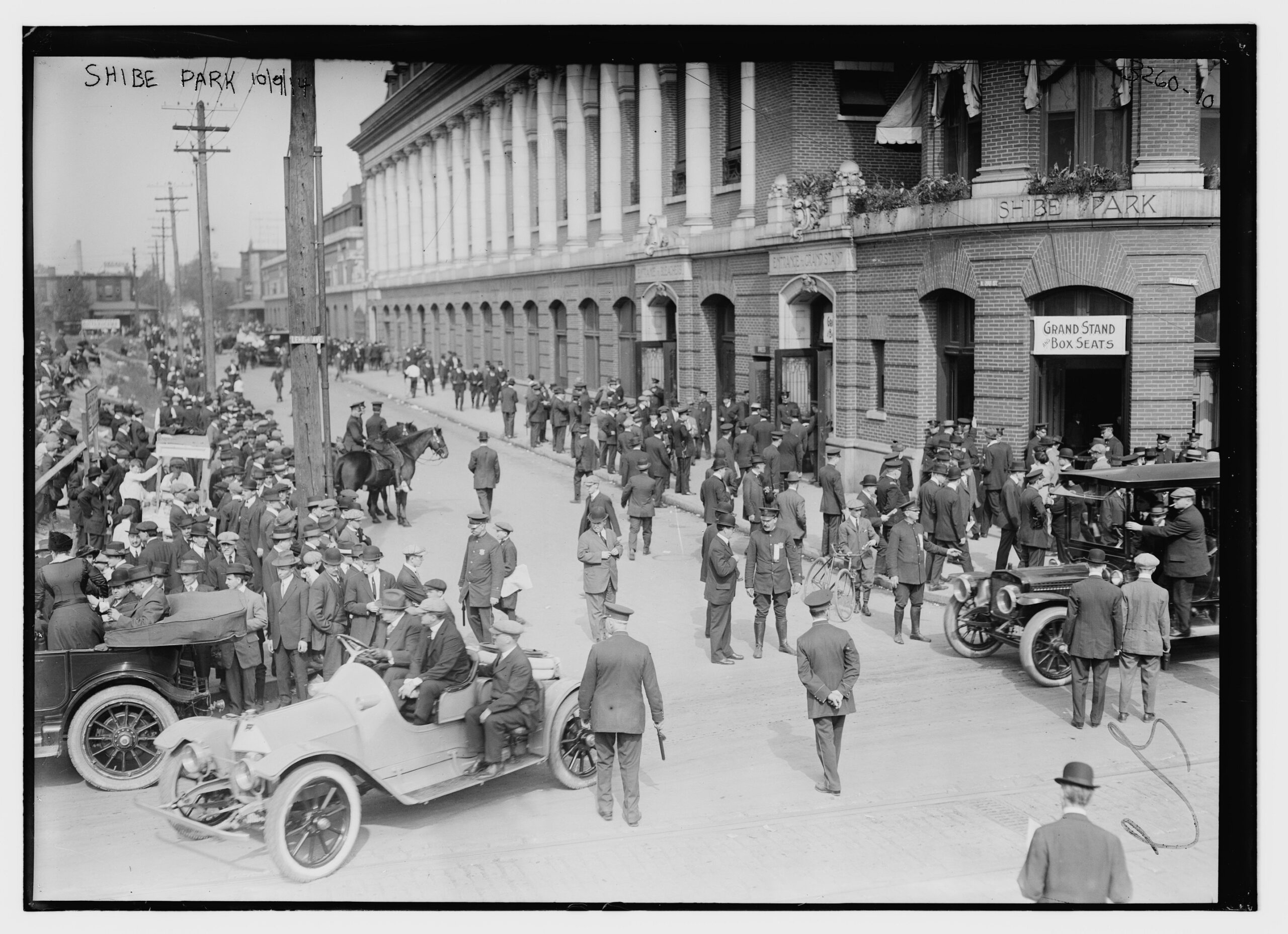 Vintage photograph showing large group of people outside brick building standing or sitting in cars.