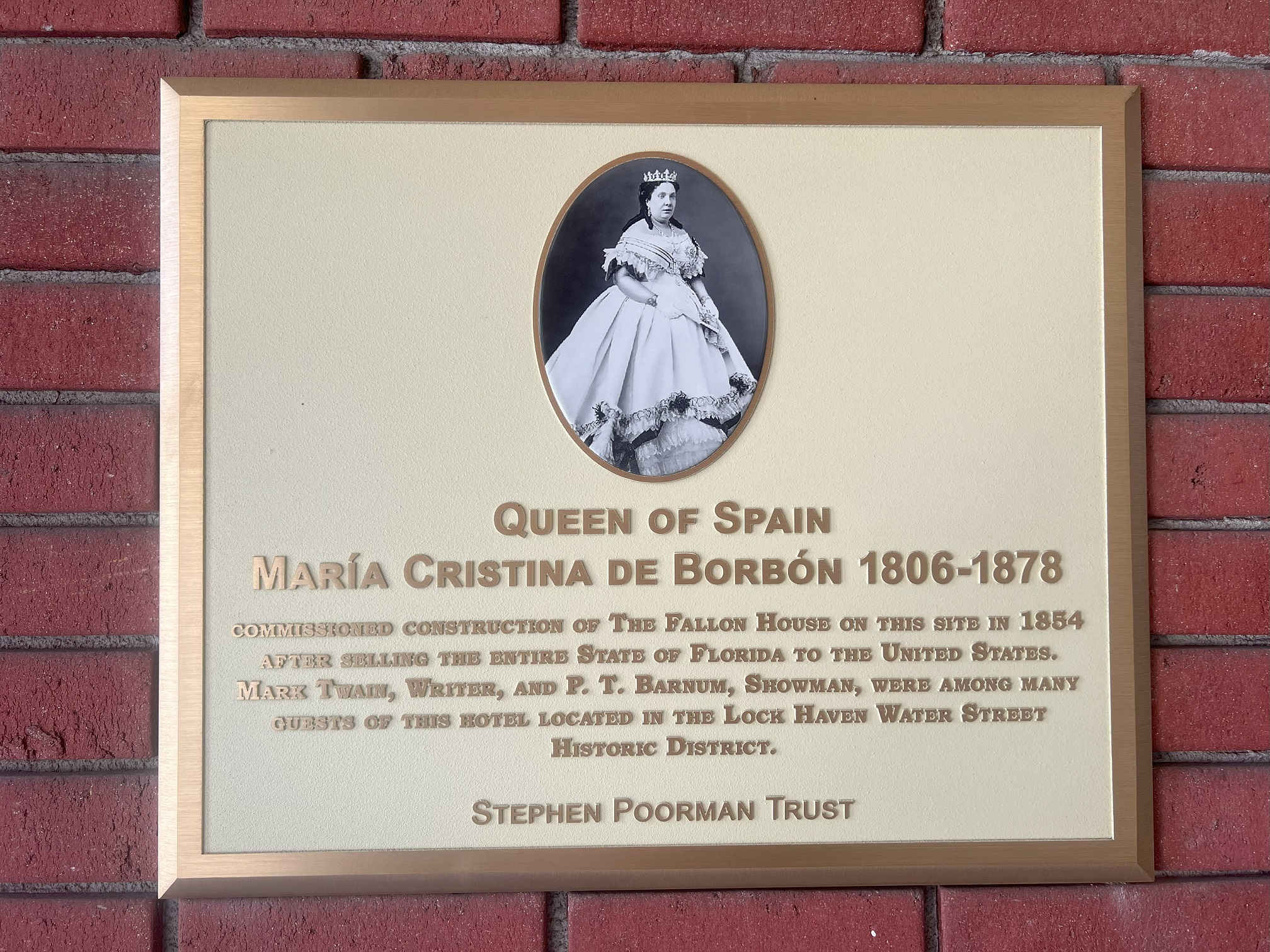 Square sign on wall with image of a woman above the words "Queen of Spain Maria Cristina de Borbon 1806-1878, Commissioned Construction of The Fallon House on this site in 1854 after selling the entire state of Florida to the United States. Mark Twain, writer, and P.T. Barnum, Showman, were among many guests of this hotel located in the Lock Haven Water Street Historic District."