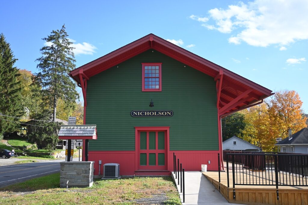 Two story wood building painted green and red with grassy front and sidewalk to large platform.