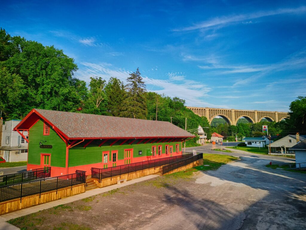 Long one-story wooden building at left side of unpaved lot with large arched bridge in background.