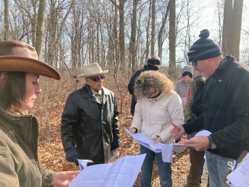 Group of people stand in wooded area looking at paper.