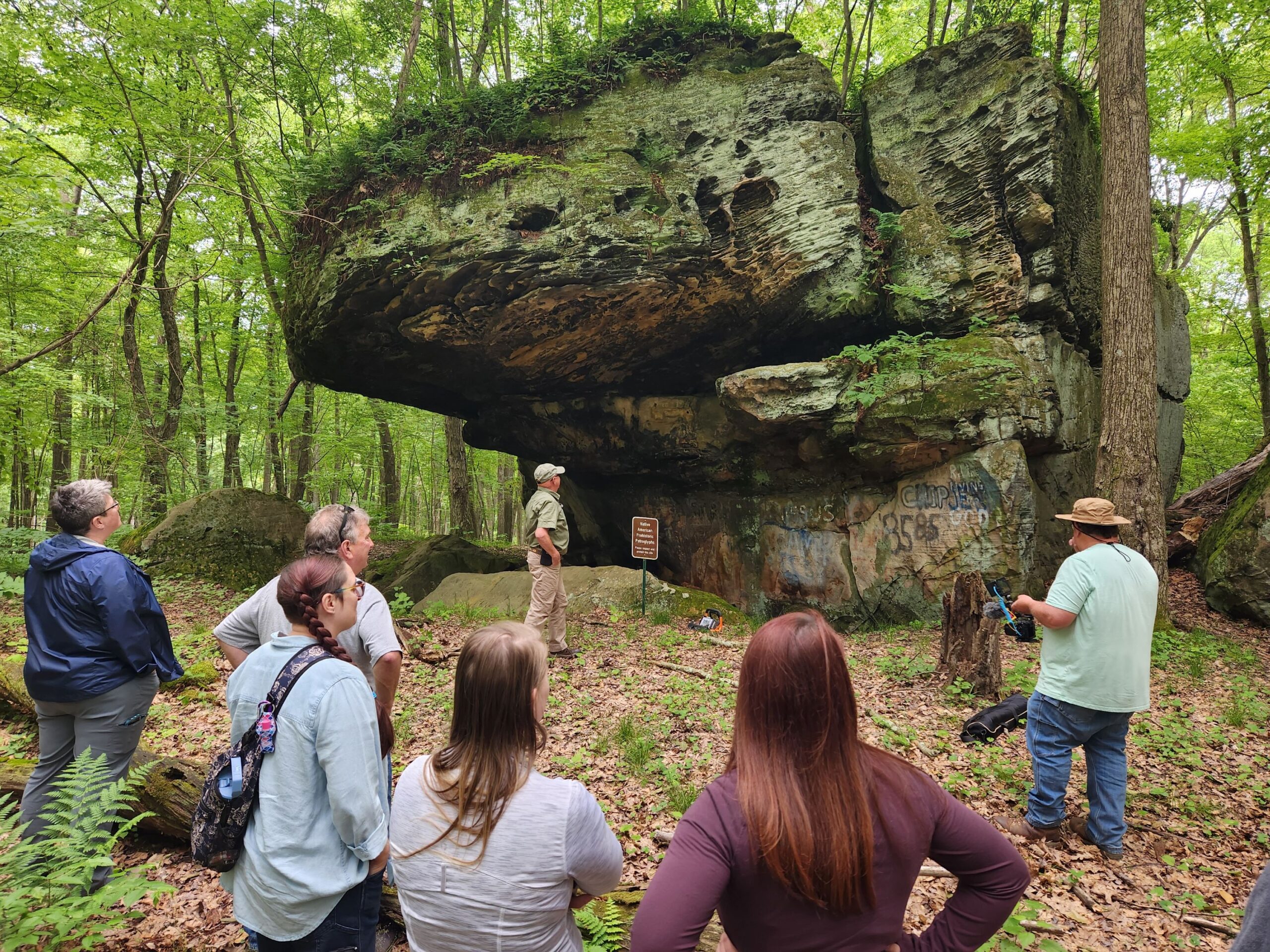 A group of people looking at a large rock formation in the woods.