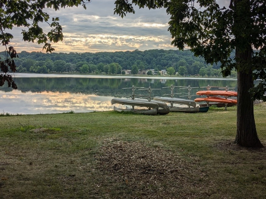 Row of canoes and kayaks on grass at the side of a large lake.