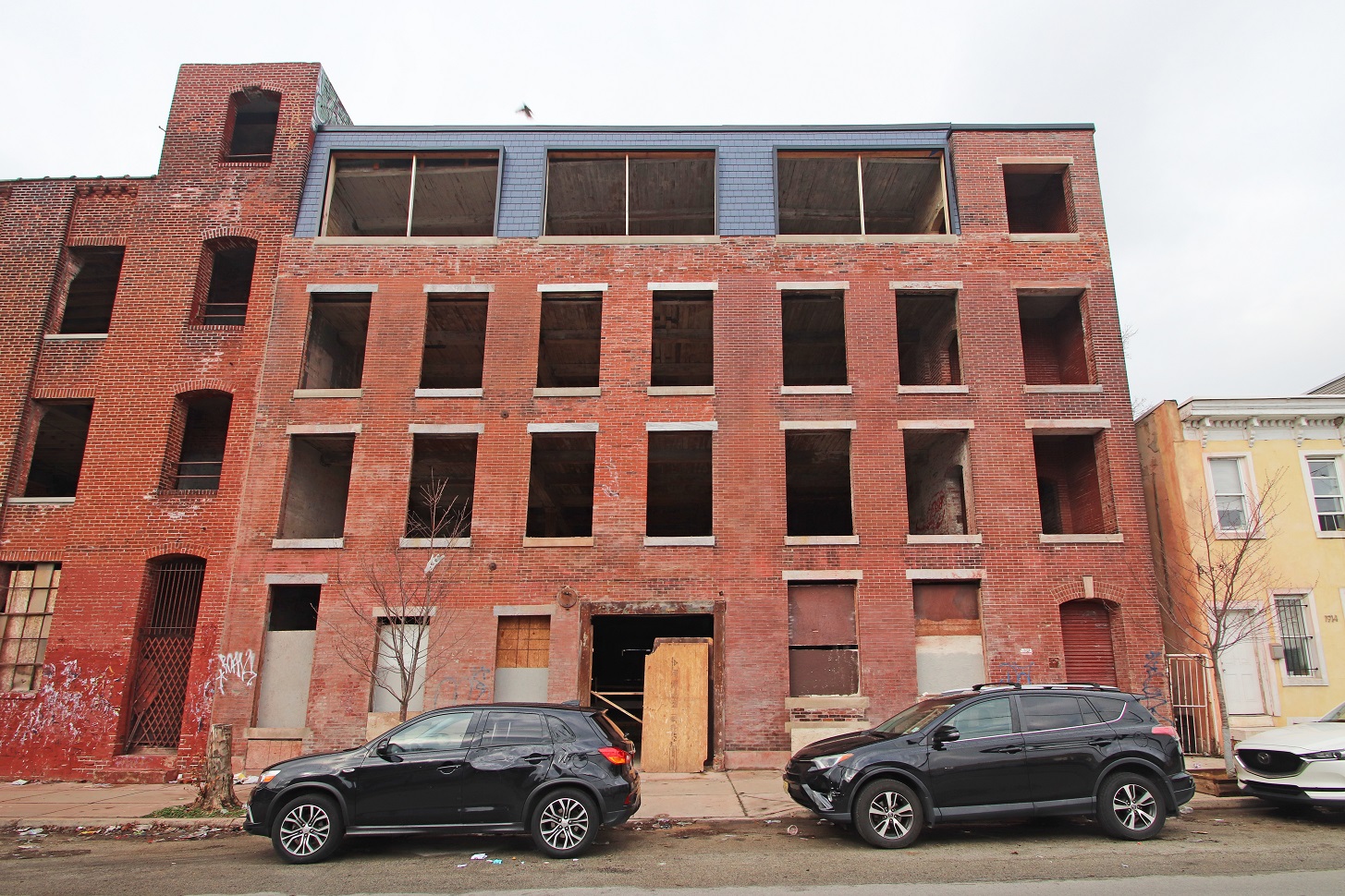 Large 4-story brick building with many empty window openings in the middle of an urban block.