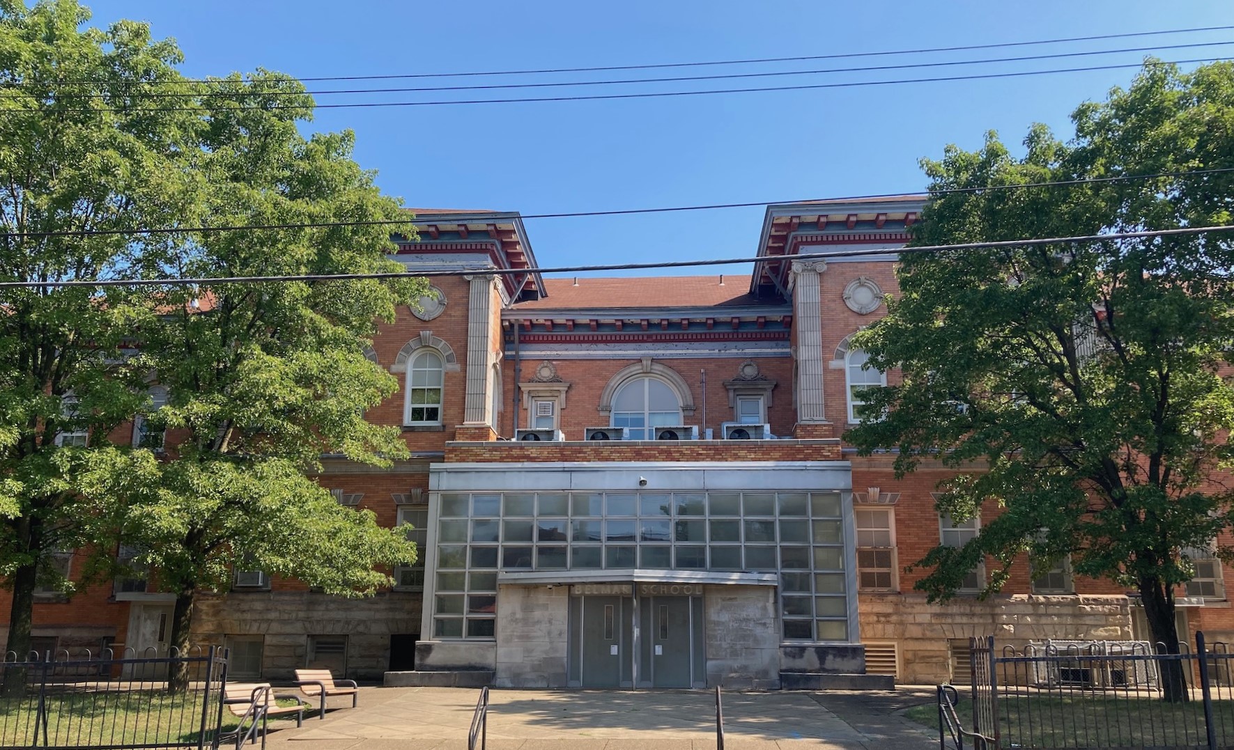 Large brick school with modern two-story entrance and large trees in front grassy areas.