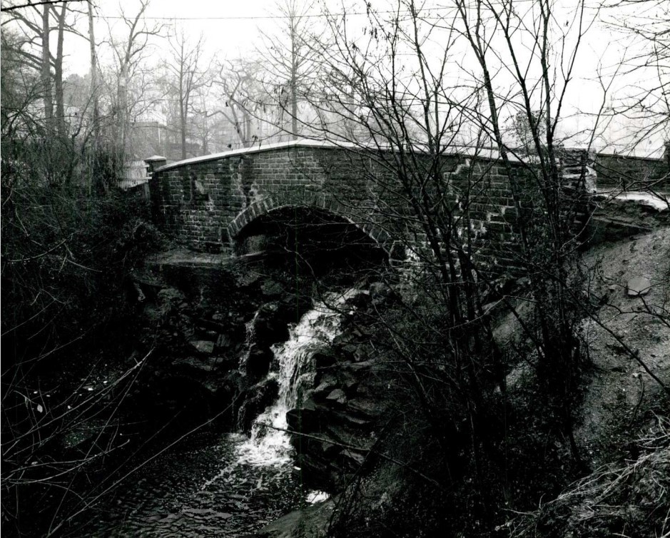 Water runs over stones below a small arched stone bridge.