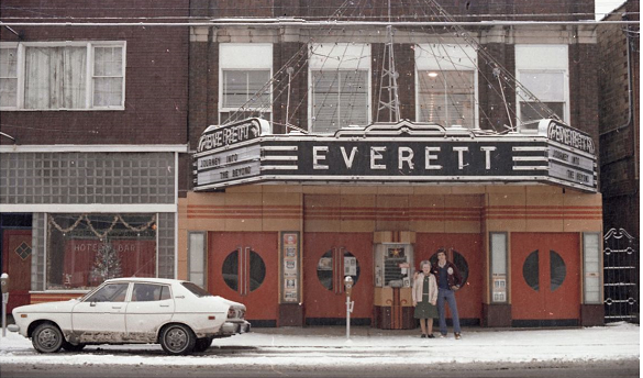 Two people stand in front of a theater adorned with Christmas lights and after a snowstorm.