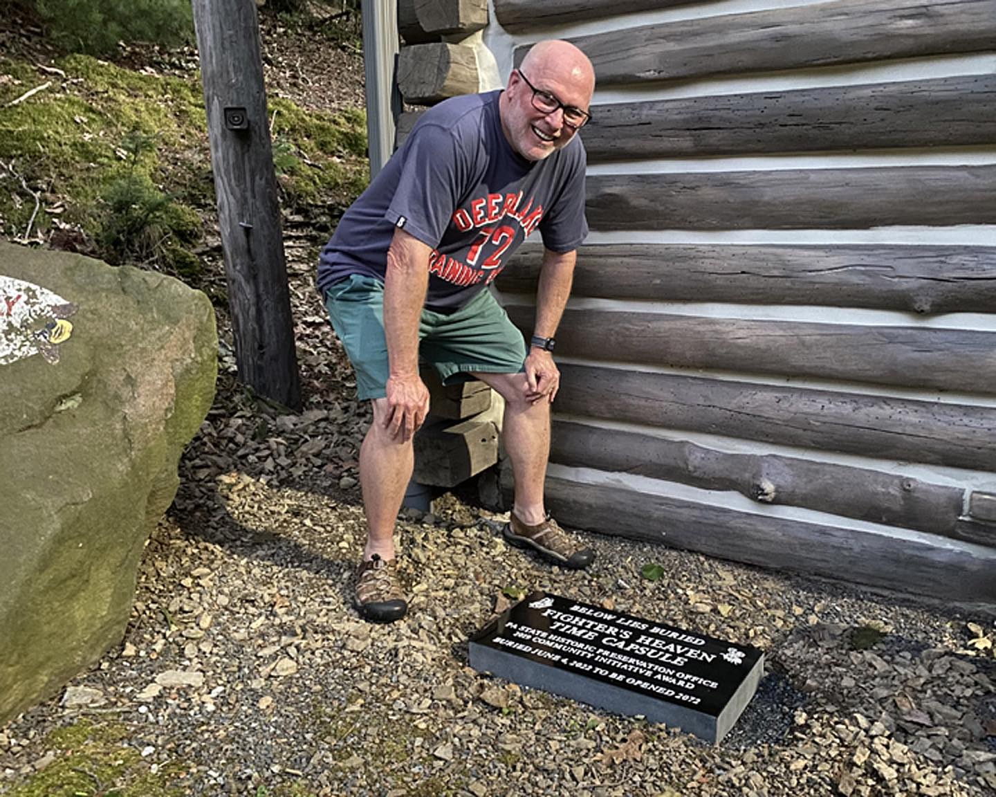 Man stands outside next to log building.