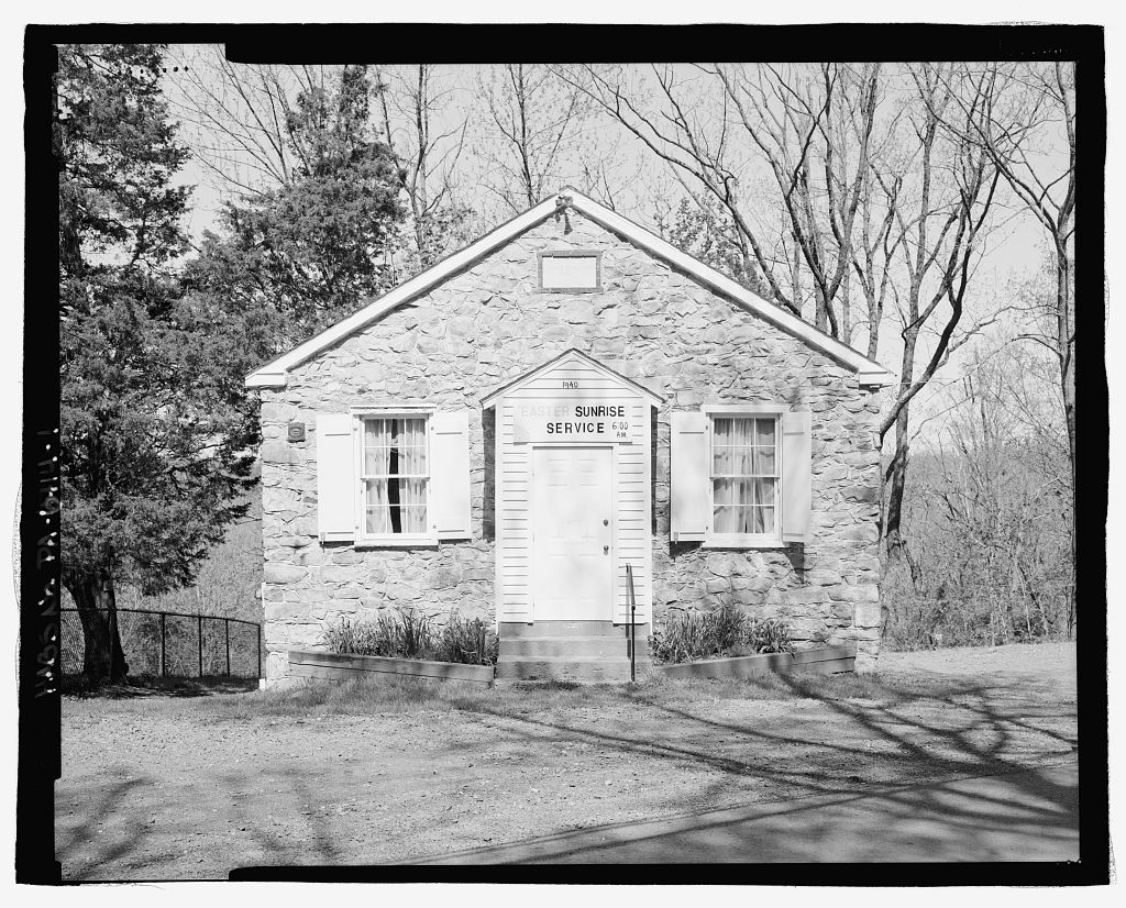 Black and white photograph of the front of a small one-story stone building with a gable roof surrounded by dirt and woodland.