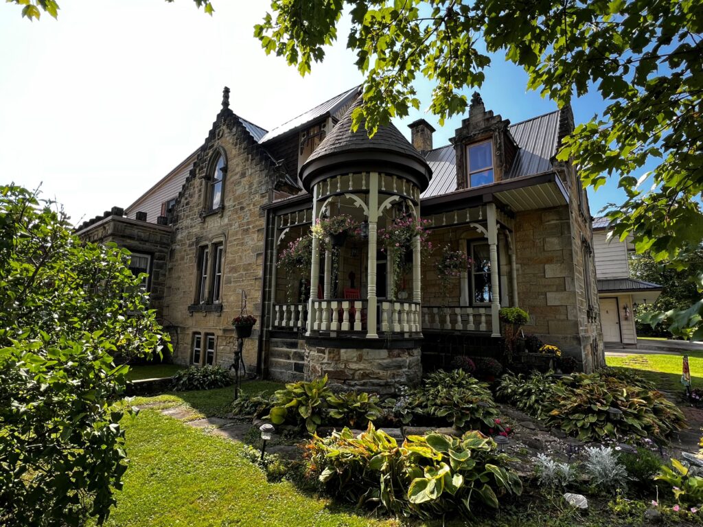 Small one-story stone house with wood porch and surrounded by lawns and gardens.